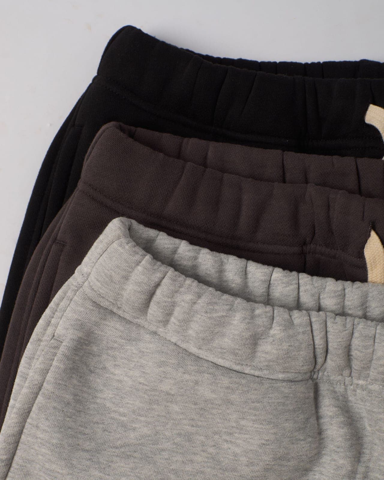 Three pairs of folded joggers in grey, brown and black, stacked to show the elasticated waistbands